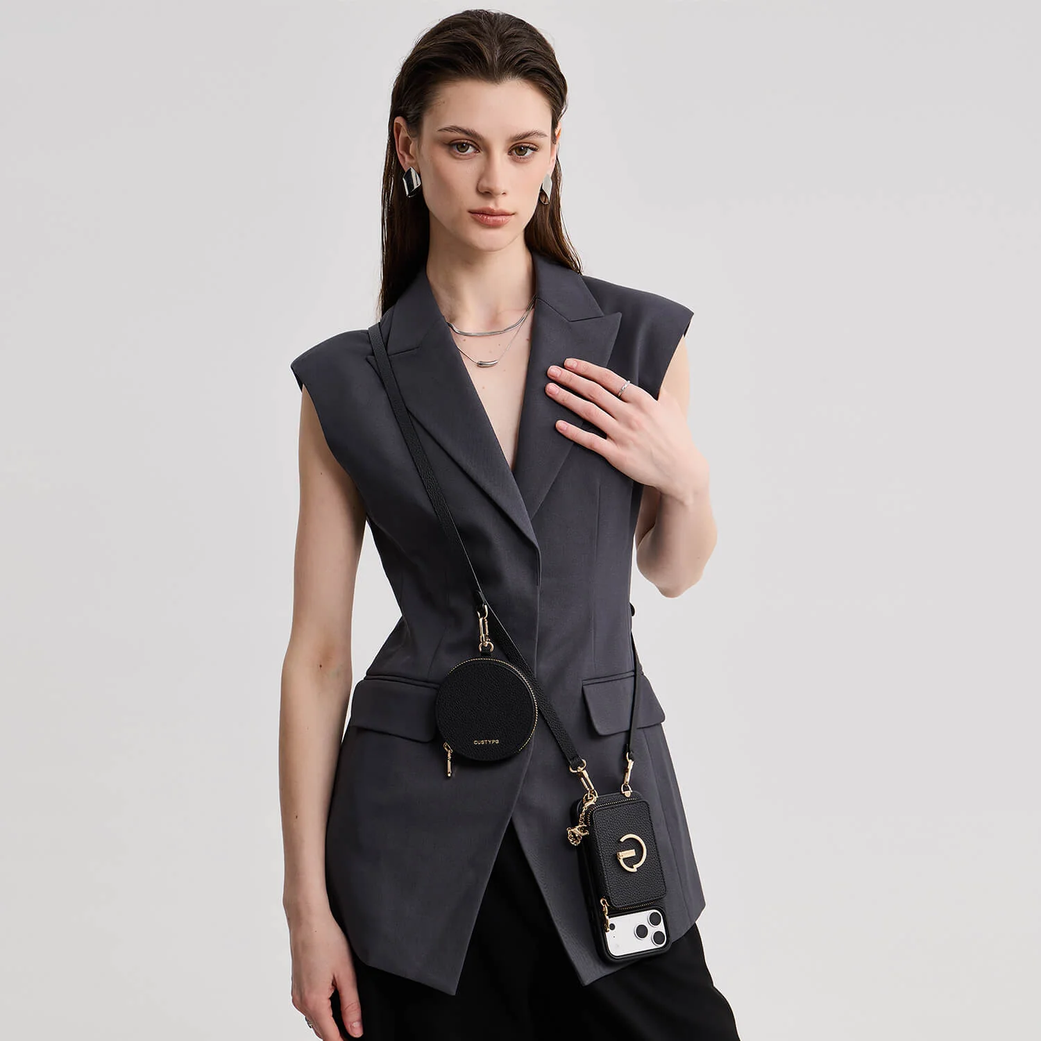 Model in a dark sleeveless blazer dress, paired with two round black crossbody bags and a phone case on one strap.